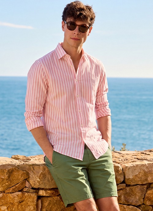 Man wearing a pink striped linen and cotton shirt with green shorts, an elegant summer outfit by the sea